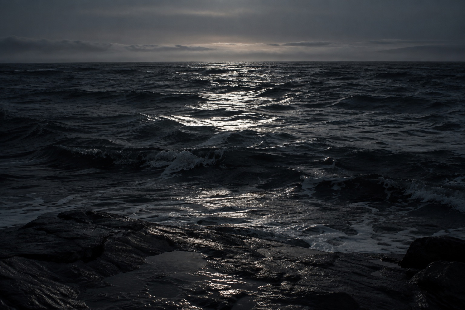 Silver light crossing dark ocean waves and wet shoreline stone.