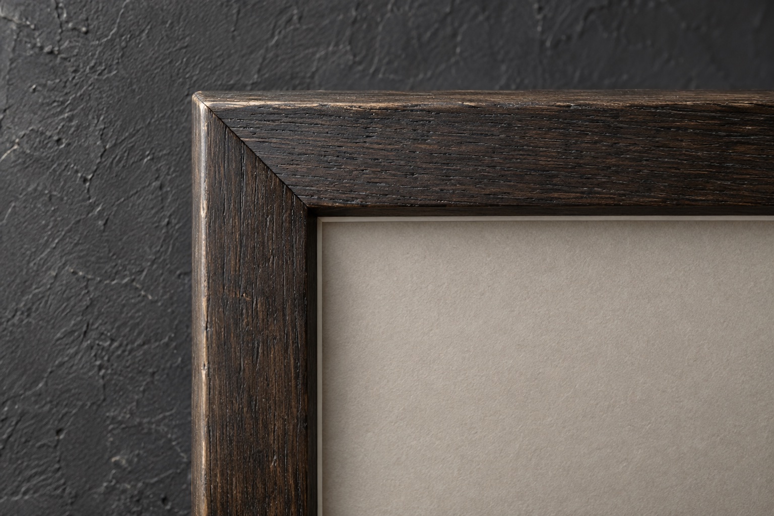The corner of a dark oak frame with a plain grey mount against textured plaster.
